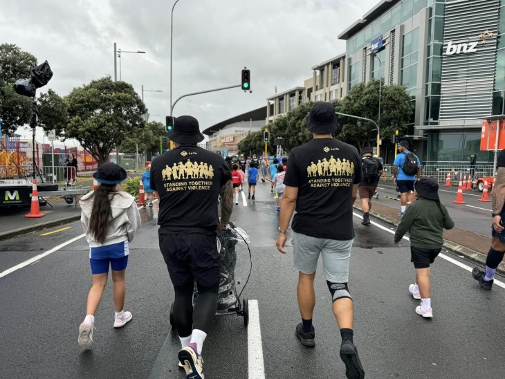 A pair of fathers from Le Va, shown from behind walk down the road at Round the Bays. On the back of their shirts is a slogan saying 'Standing together against violence'. 